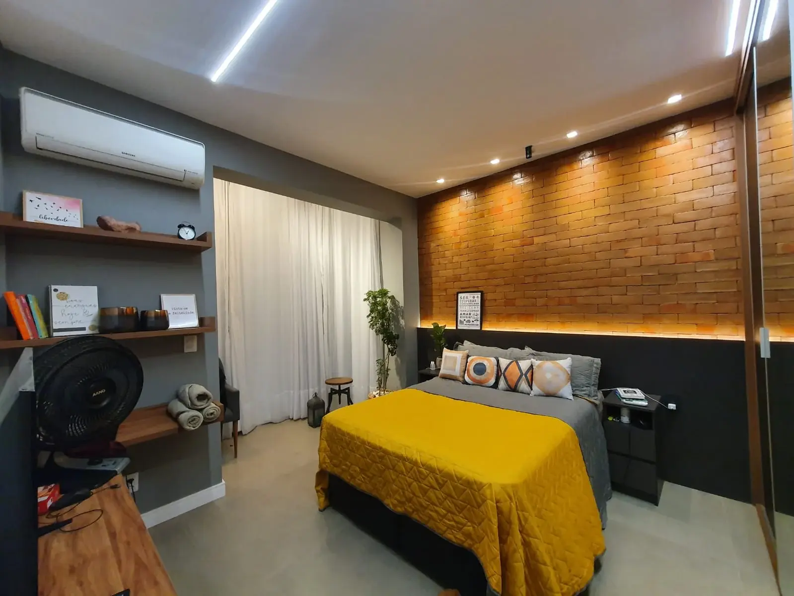 Quarto após a modernização, com parede de tijolinhos, iluminação embutida, cama com colcha amarela e prateleiras